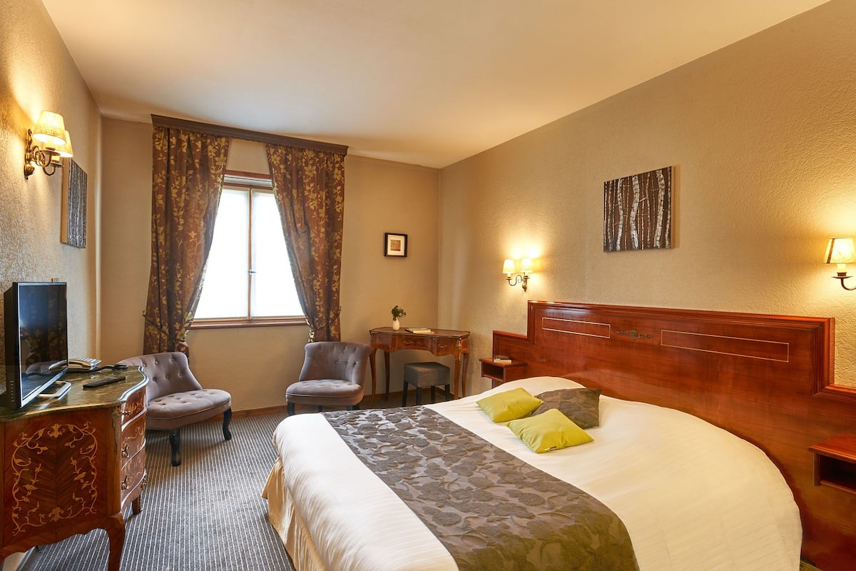 A hotel room with a large bed, a desk with a computer, a chair, a small table, and a window with curtains.