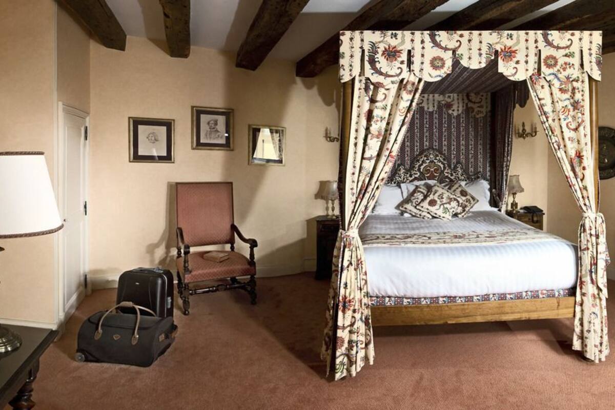 Un lit traditionnel à baldaquin, une chaise en bois, une table de chevet avec une lampe et une valise posée sur le sol.