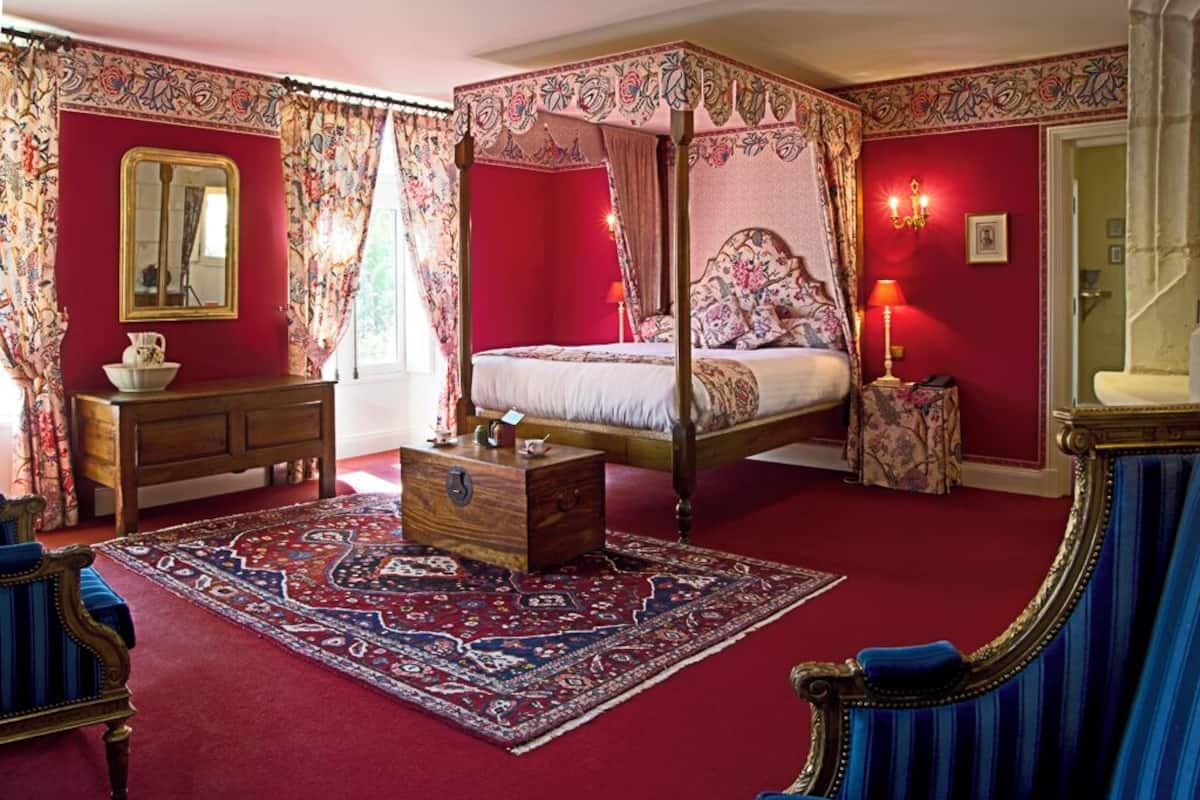 Un lit à baldaquin, une commode en bois, une chaise assortie et un tapis à motifs dans une pièce aux murs rouges et dotée d’un grand miroir.