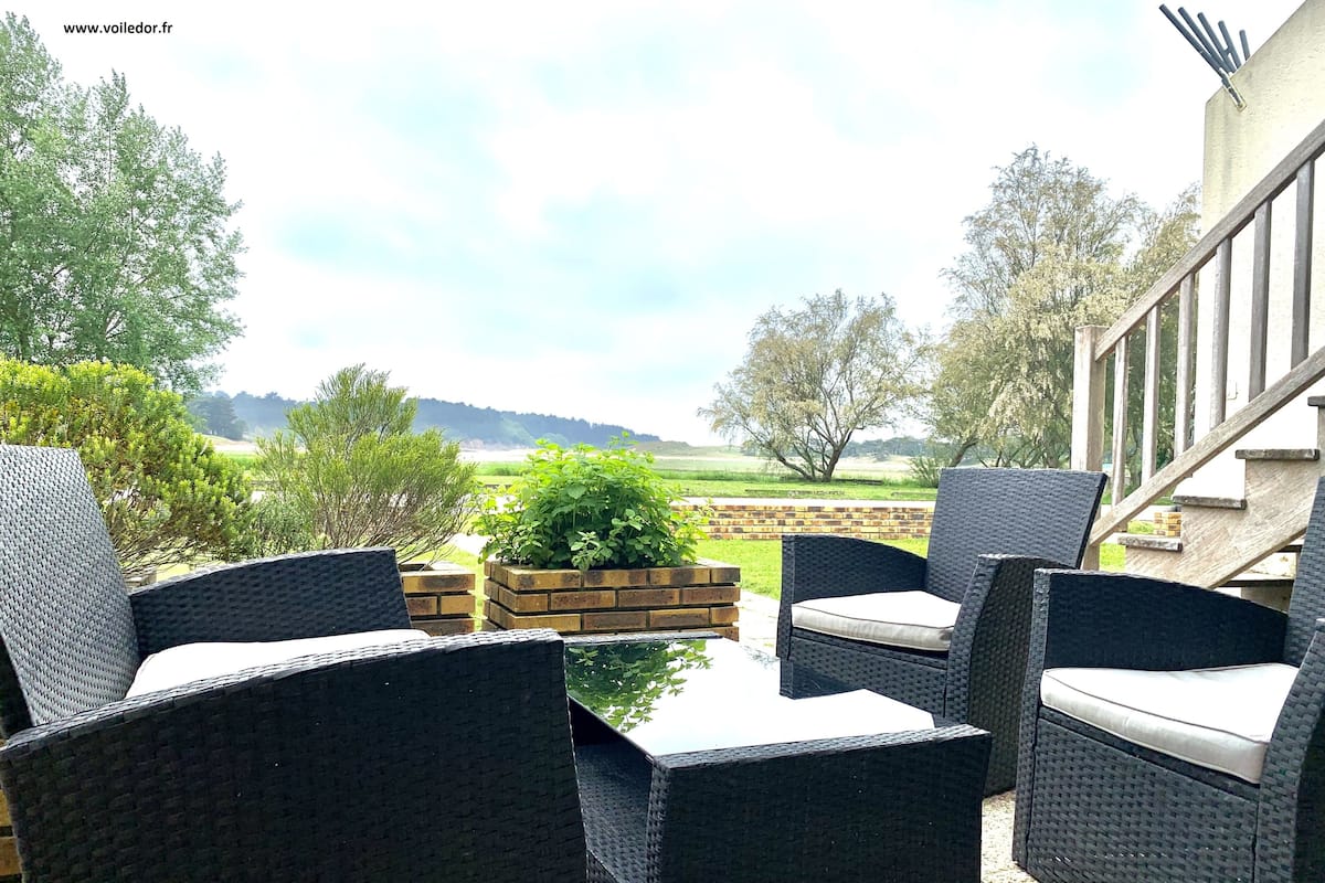 A patio with wicker furniture and a glass table, overlooking a grassy field and trees.