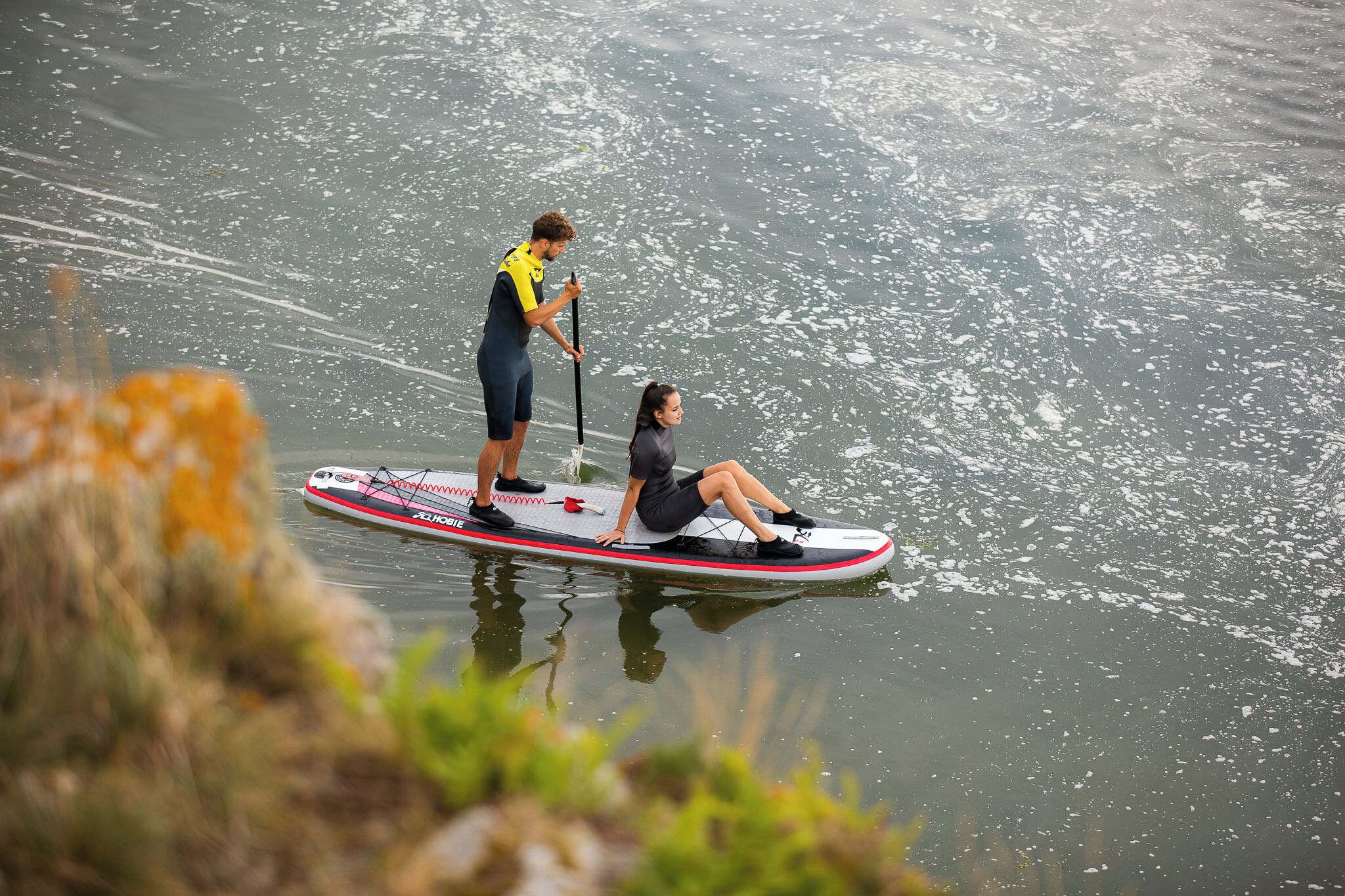 A person sitting on a paddleboard in a body of water, with another person standing and holding a paddle.