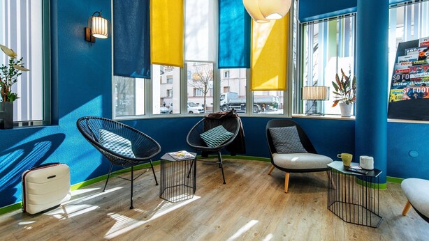 A room with a blue and yellow color scheme, featuring a large window, two black wicker chairs, a small table, and a suitcase on the floor. - ibis Styles Lyon Croix Rousse (Lyon)