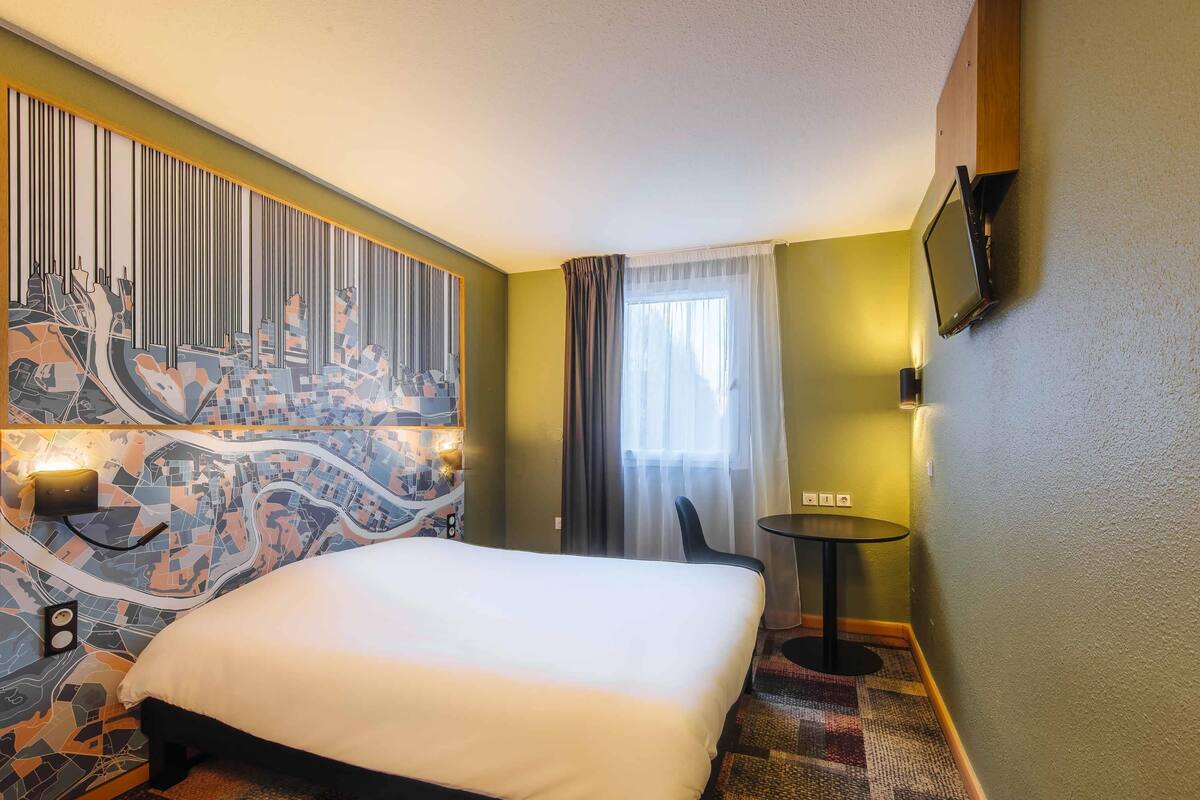 A hotel room with a large bed, a cityscape mural on the wall, a small table, and a TV mounted on the wall.