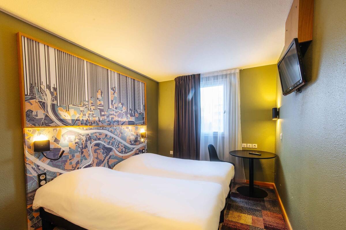 A hotel room with a large bed, a small table, a TV mounted on the wall, and a cityscape mural on the wall.
