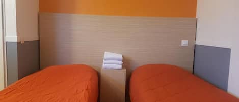 Hotellihuoneessa on kaksi sänkyä, joissa on molemmissa oranssi sängynpeite. Sängyissä on puinen pääty ja niiden vieressä pieni puinen yöpöytä, jonka päällä on siististi taiteltuja pyyhkeitä.