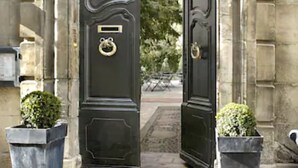 A black door with a brass knocker and a small plaque, flanked by two square planters with green shrubs. - L'Hôtel Particulier (Arles)