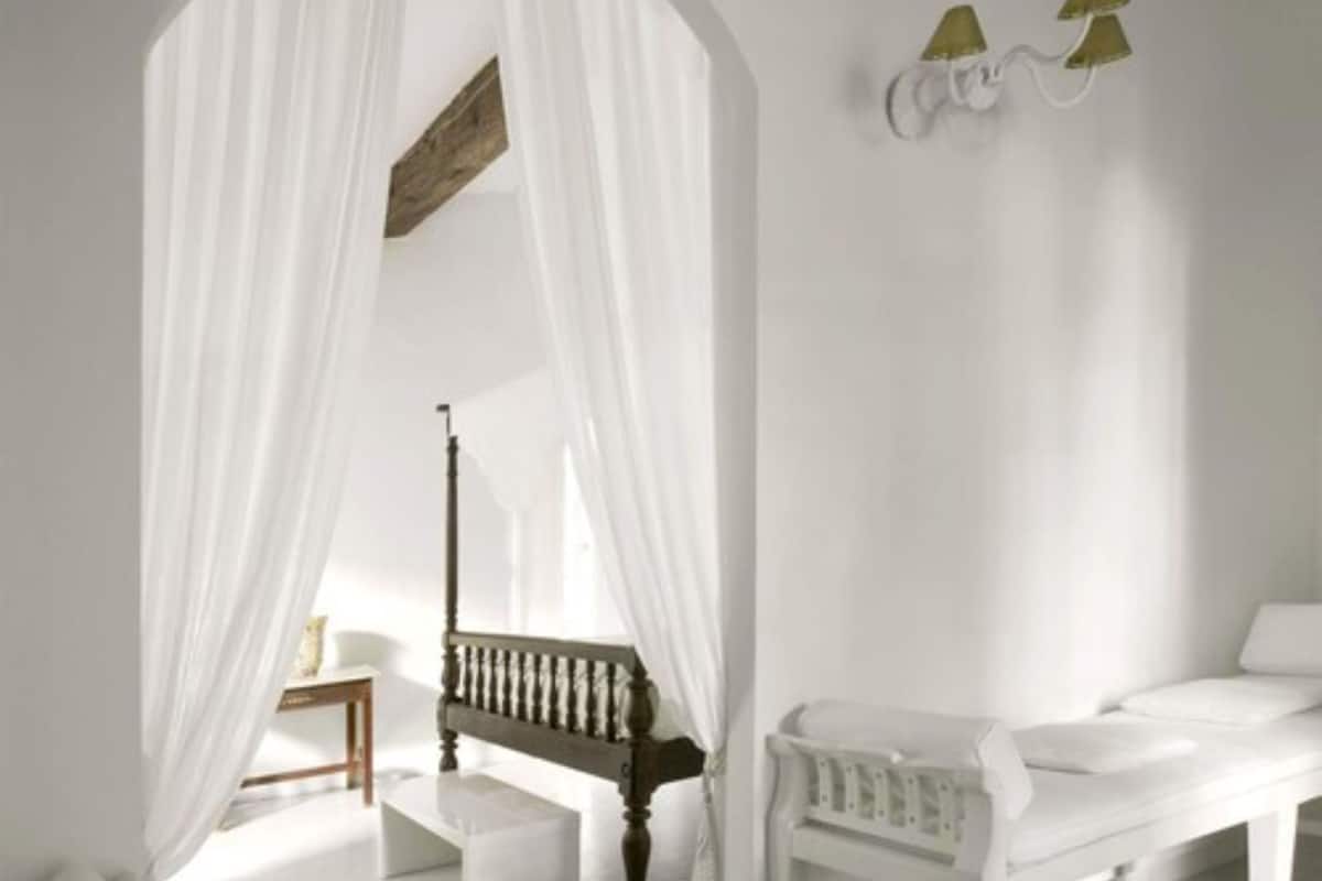 A room with a white bed, a wooden bench, a small table, and a wall-mounted lamp.