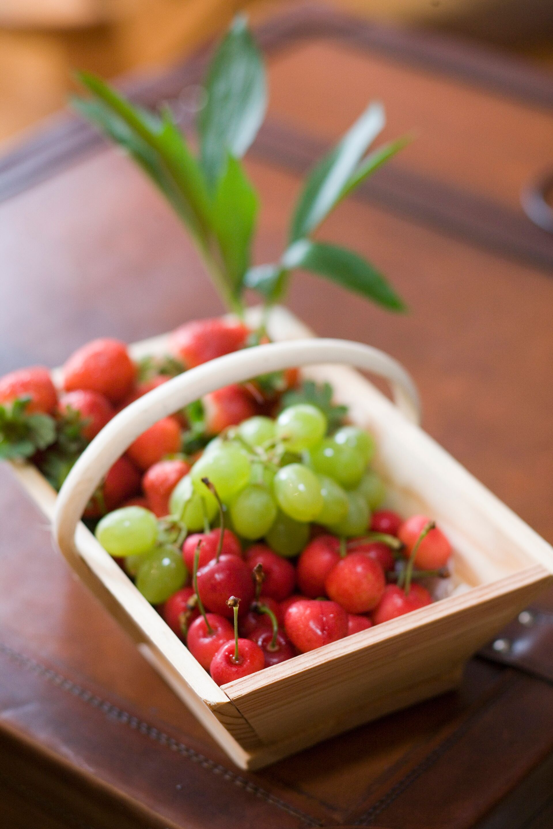 A wooden fruit basket containing red and green grapes and cherries on a wooden table.