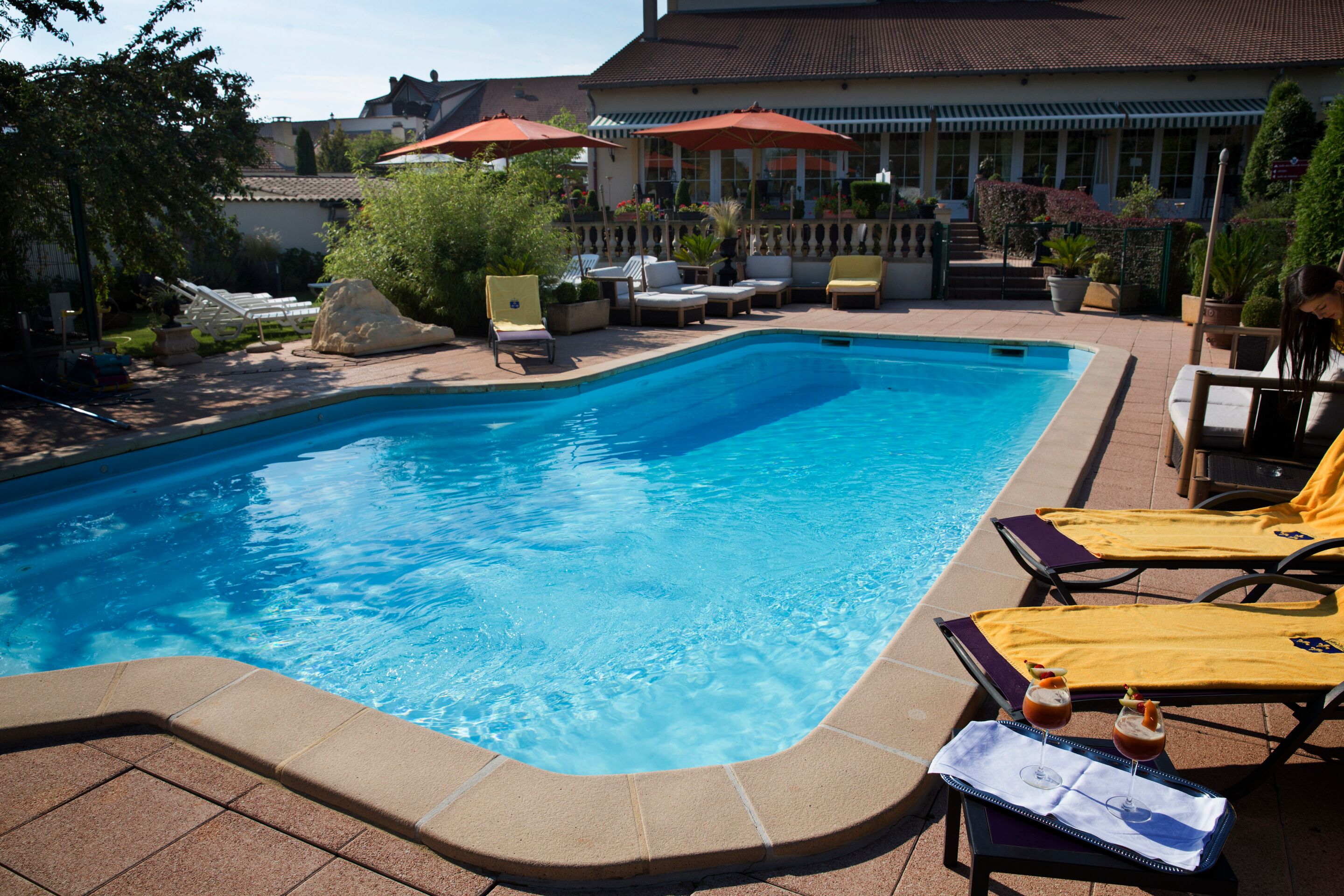 A swimming pool with lounge chairs and umbrellas, surrounded by potted plants and a building in the background.