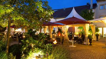 An outdoor dining area with tables and chairs, surrounded by greenery and illuminated by warm lighting.