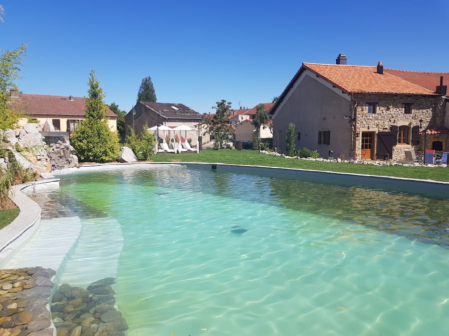 A clear swimming pool with a stone border, surrounded by houses with red-tiled roofs and stone walls.