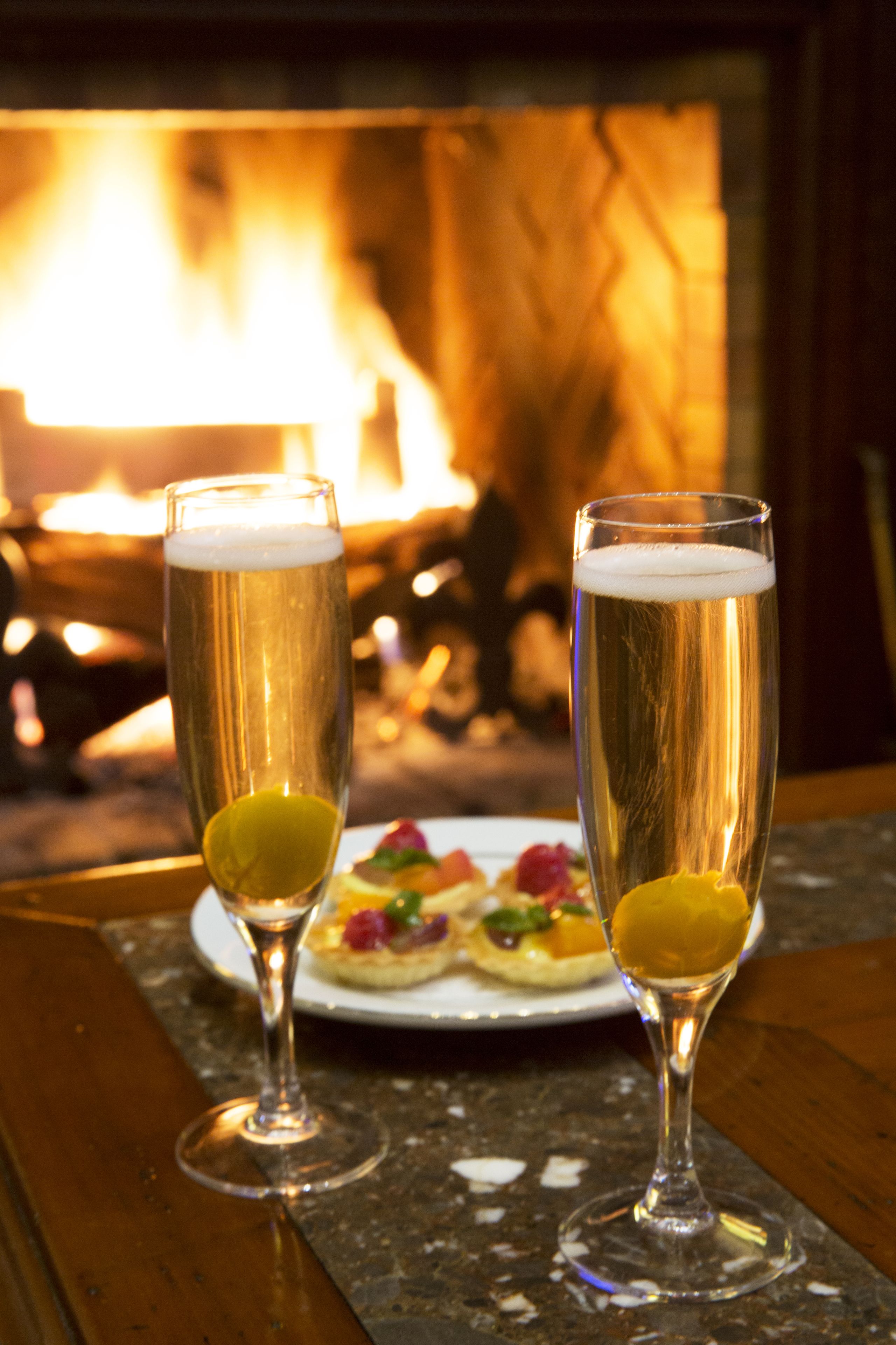 Dos vasos de cerveza con rodajas de limón sobre una mesa, con un plato de comida y una chimenea al fondo.