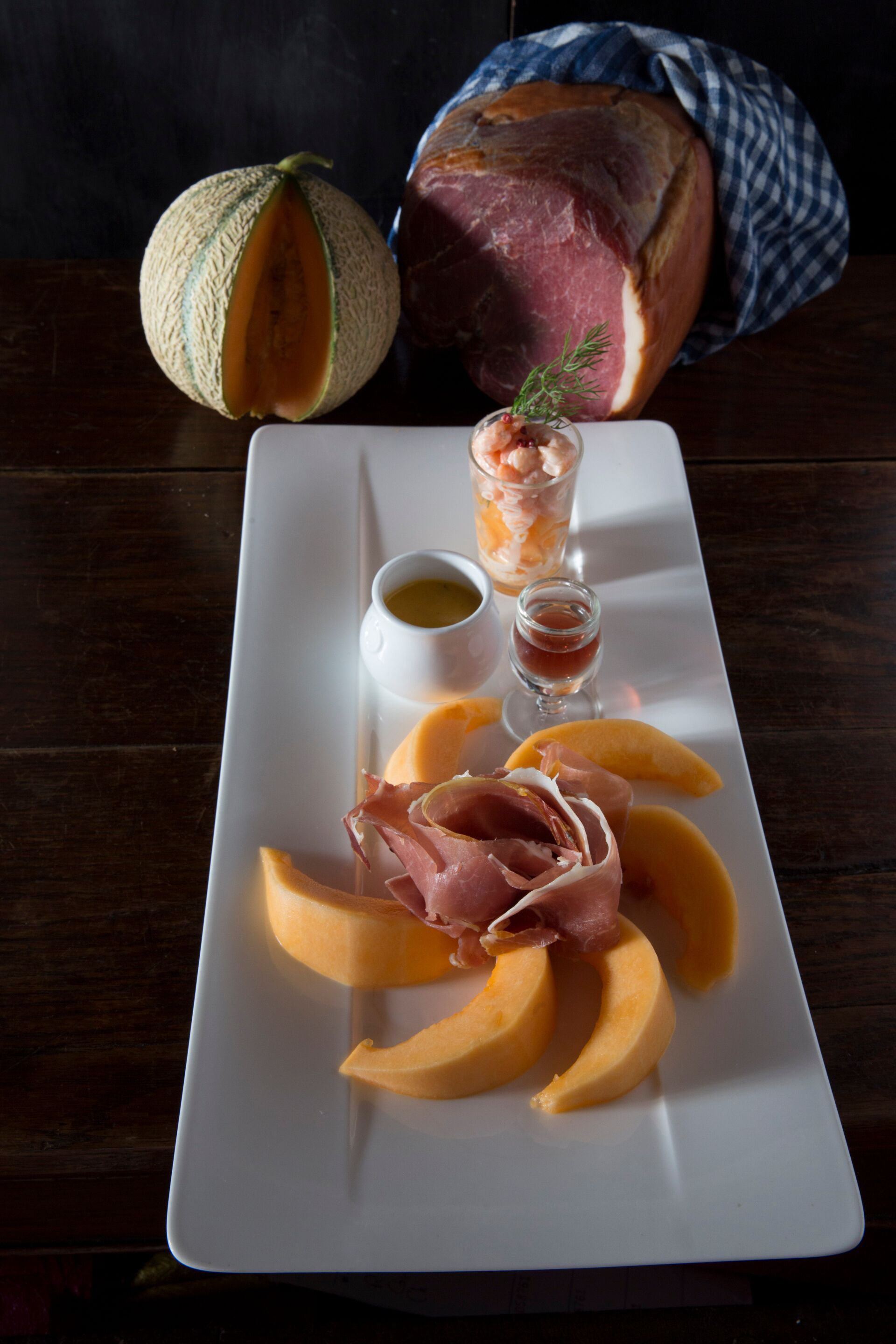 Un plato rectangular blanco con melón en rodajas, jamón serrano y dos pequeños vasos de salsa sobre una mesa de madera.