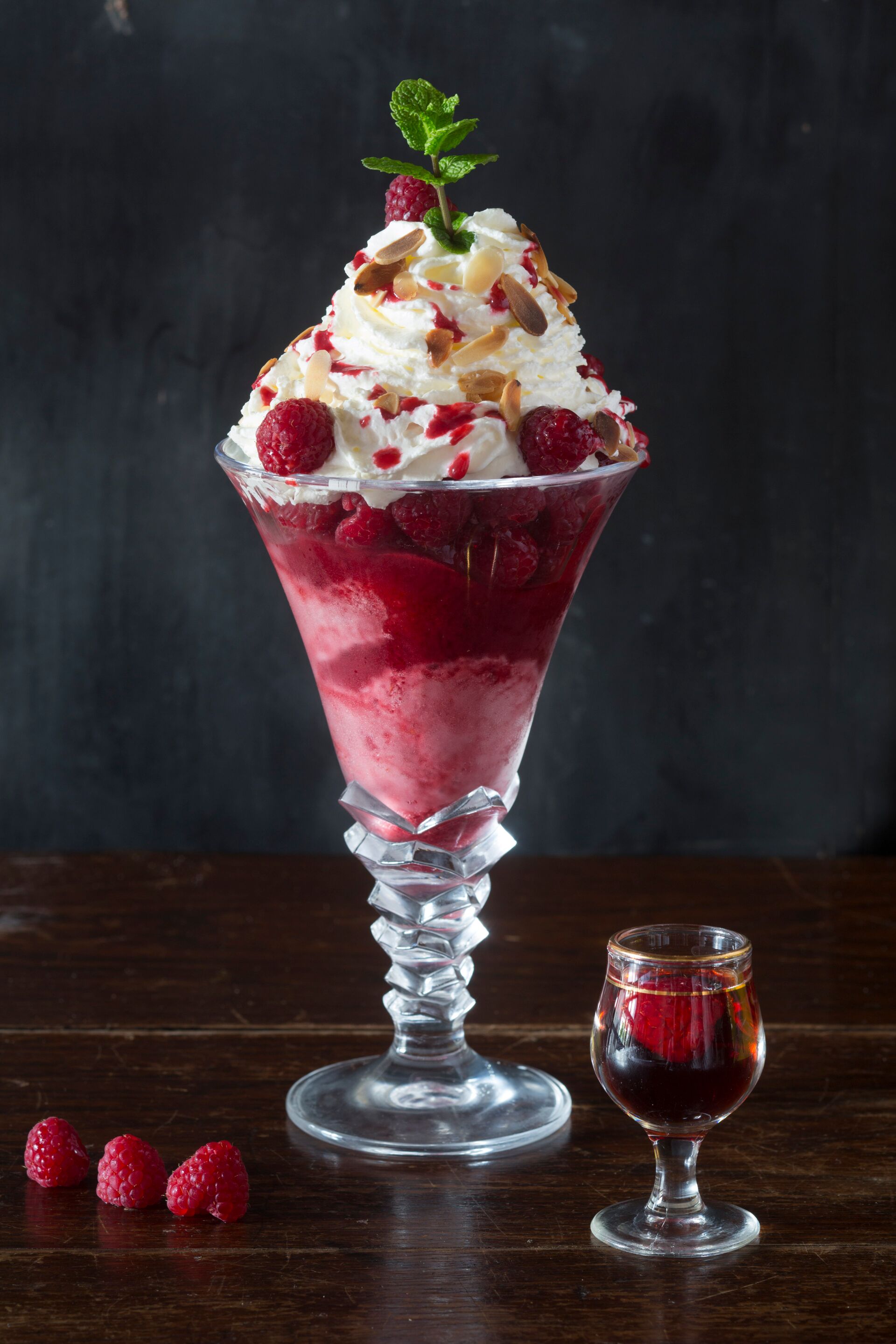 Un dessert servi dans un verre à pied, accompagné d’un plus petit verre et de framboises disposées sur une surface sombre.