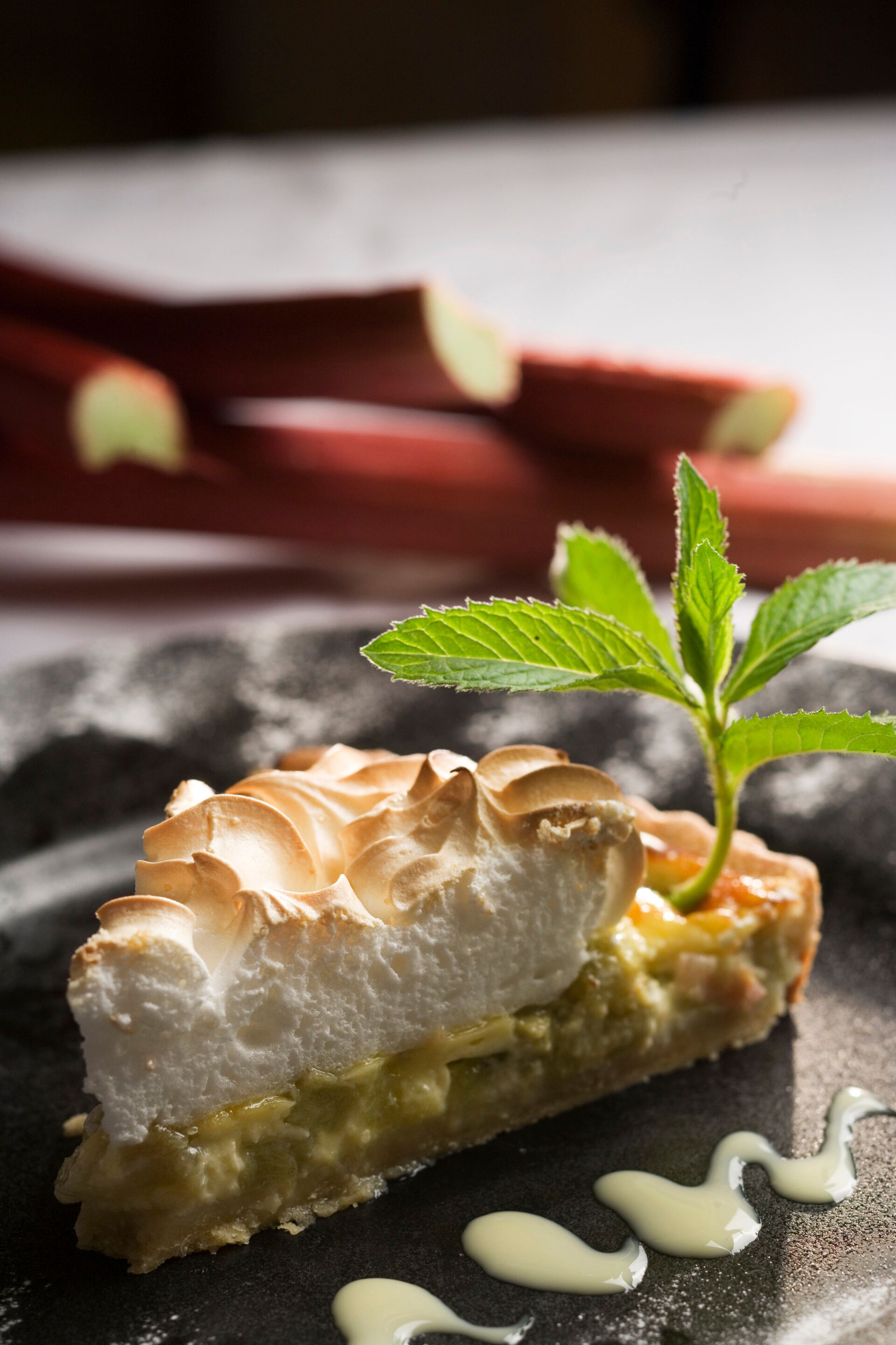 Un postre con cobertura de merengue, una hoja de menta como adorno y un hilo de salsa sobre un plato oscuro.