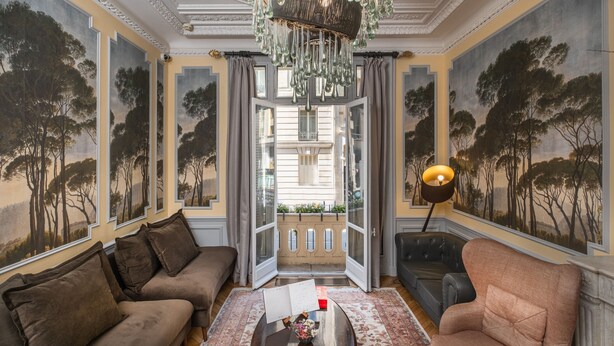 A room with a large window, a chandelier, and a view of a building outside. - Hôtel Le Paris Singulier (Paris)