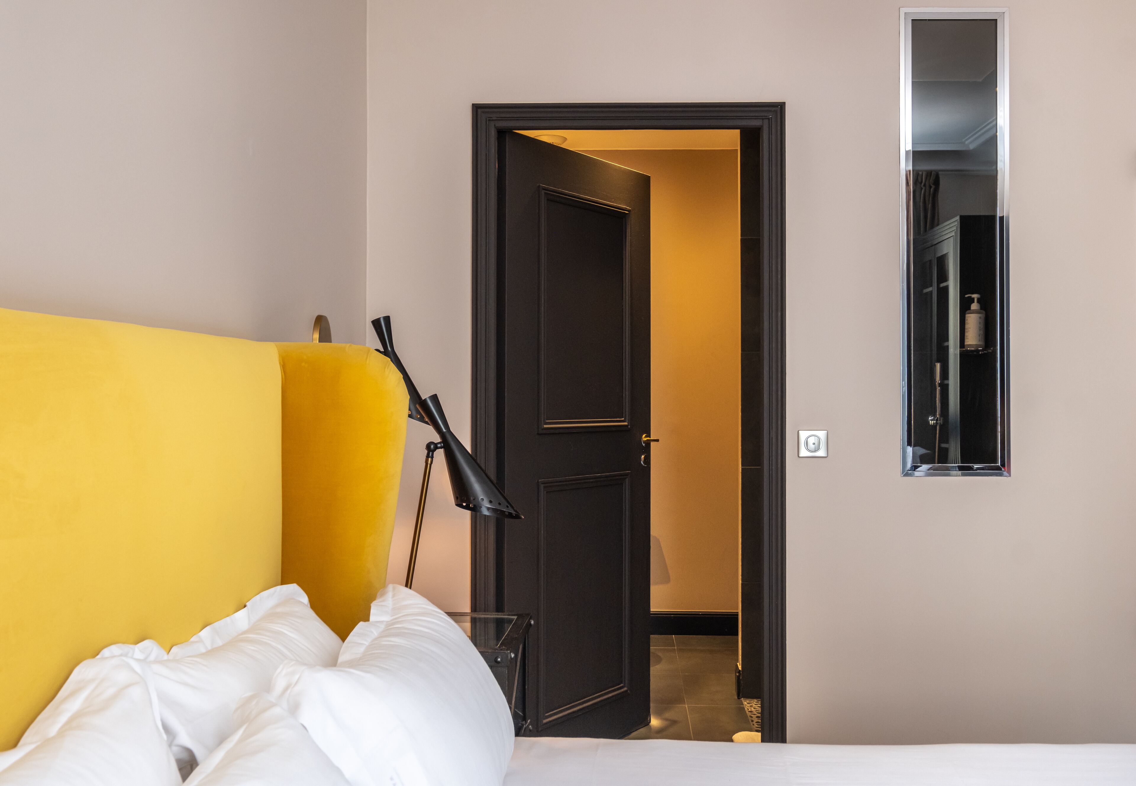 A hotel room with a bed, a lamp, a door, and a mirror.