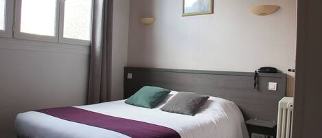 Una habitación de hotel con una cama, una mesita de noche, una lámpara y un ventanal con cortinas.