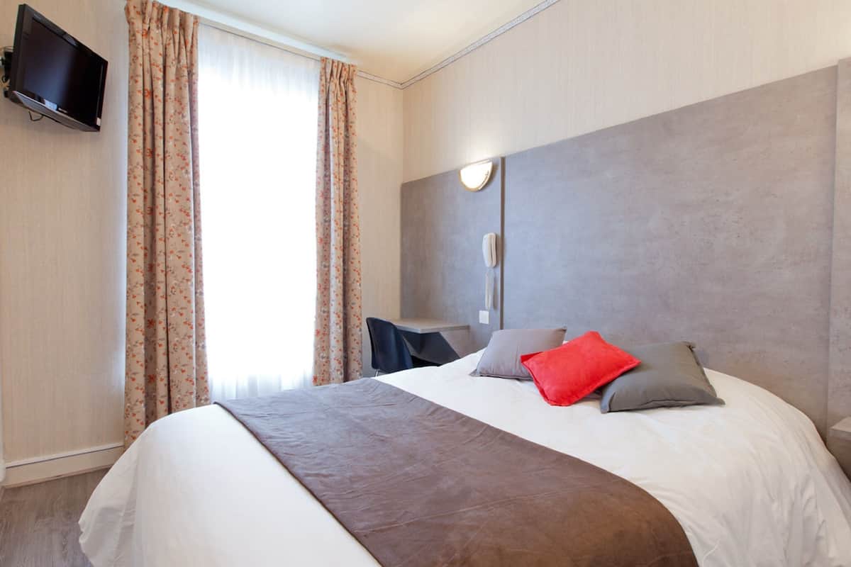 A hotel room with a large bed, a flat-screen TV mounted on the wall, a desk with a chair, and a window with curtains.
