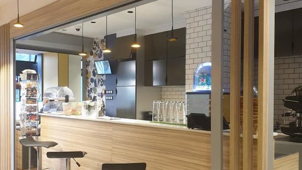 A modern bar area with wooden accents, bar stools, and a visible kitchen in the background. - Panam Hotel (Paris)