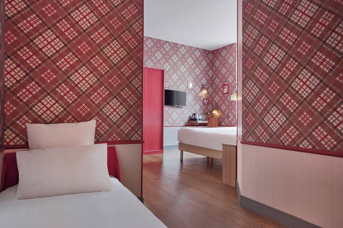 Una habitación de hotel con un estampado de cuadros en la pared, una cama con ropa blanca, una mesita de noche de madera, un televisor montado en la pared y una pequeña mesa con una lámpara y objetos decorativos.