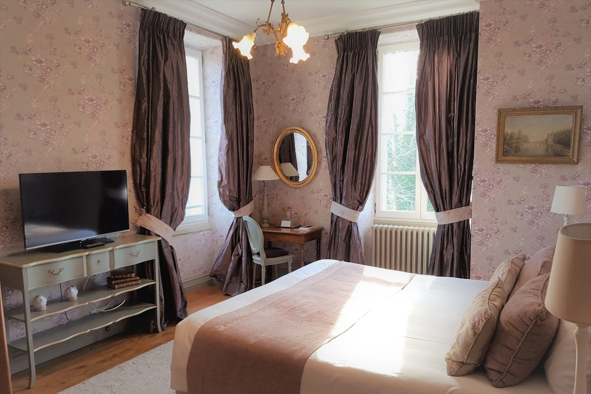Ein Schlafzimmer mit einem Bett, einem Fernseher auf einer Kommode, einem Spiegel und einem Fenster mit Vorhängen.