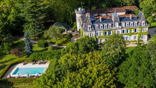 An aerial view of a large, historic mansion with a prominent tower, surrounded by lush greenery and a swimming pool.