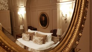 A bedroom with a large ornate mirror reflecting a bed with multiple pillows, a nightstand with books, and wall sconces.