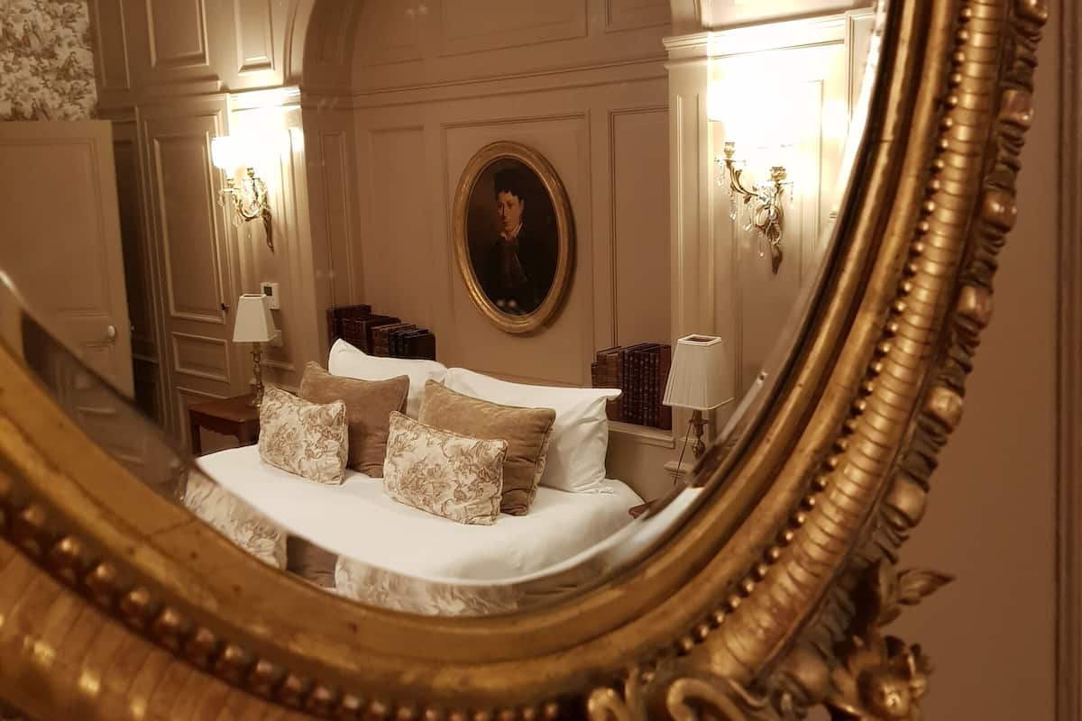 Ein Schlafzimmer mit einem großen, verzierten Spiegel, der ein Bett mit vielen Kissen, einen Nachttisch mit Büchern und Wandleuchten reflektiert.