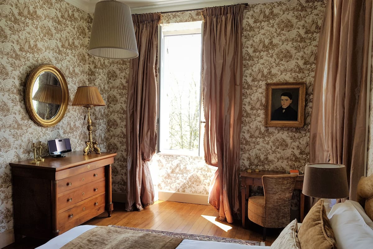 Ein Schlafzimmer mit einem Holzkasten, einem Nachttisch mit Lampe, einem Bett mit Kissen, einem Sessel und einem Fenster mit Vorhängen.