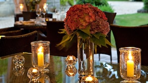 A dining table with candles, a vase of flowers, and a view of a garden and building in the background.
