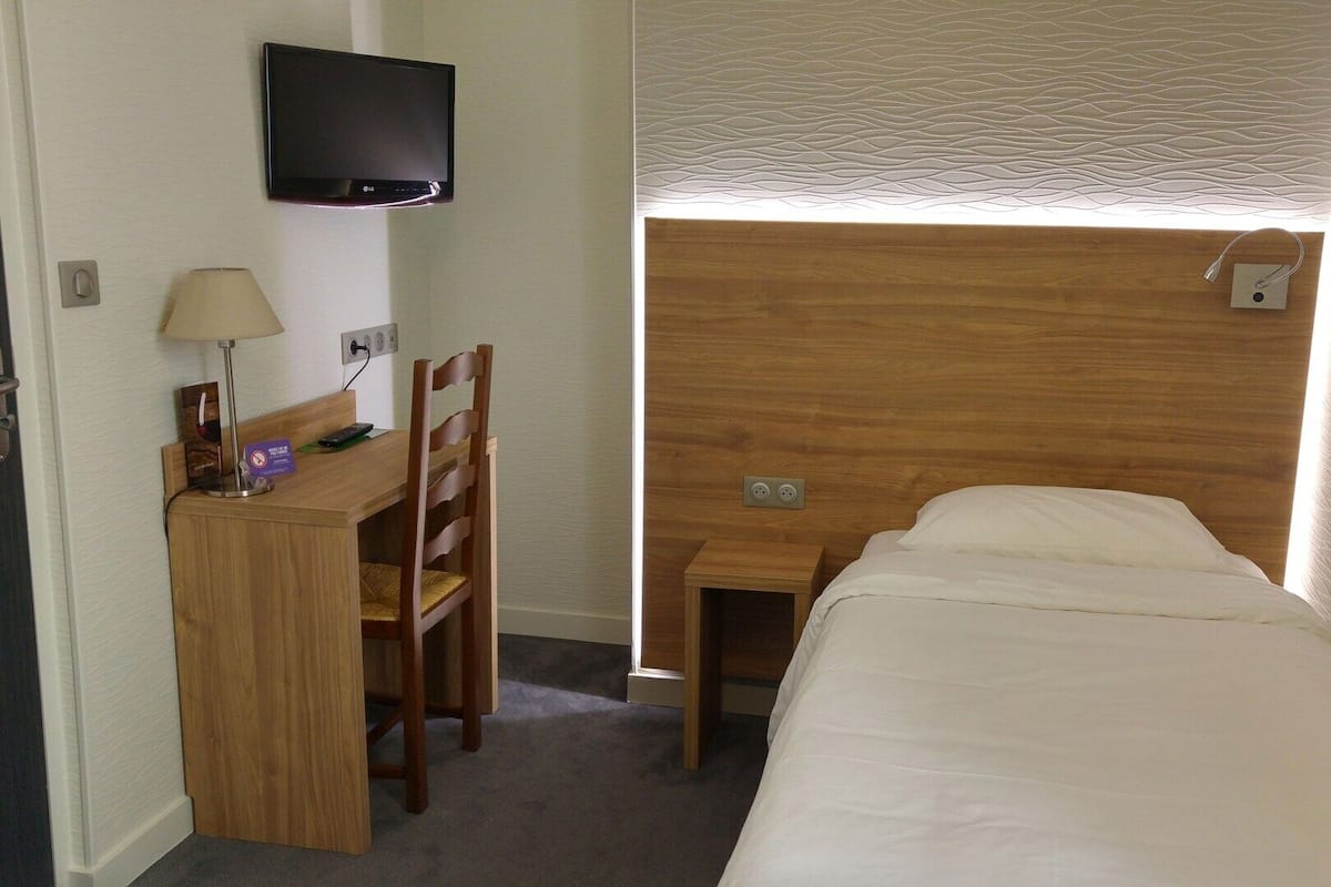 Habitación de hotel con cama, escritorio, silla, televisor y ventana con persiana texturizada.