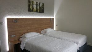 Habitación de hotel con cabecera de madera, dos camas individuales, una lámpara de pared y un cuadro con motivos forestales sobre la cabecera.