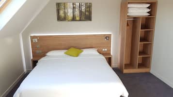 Un dormitorio con cabecera de madera, cama con ropa de cama blanca y almohada amarilla, mesita de noche, armario de madera con ropa de cama doblada y un cuadro con motivos forestales en la pared.