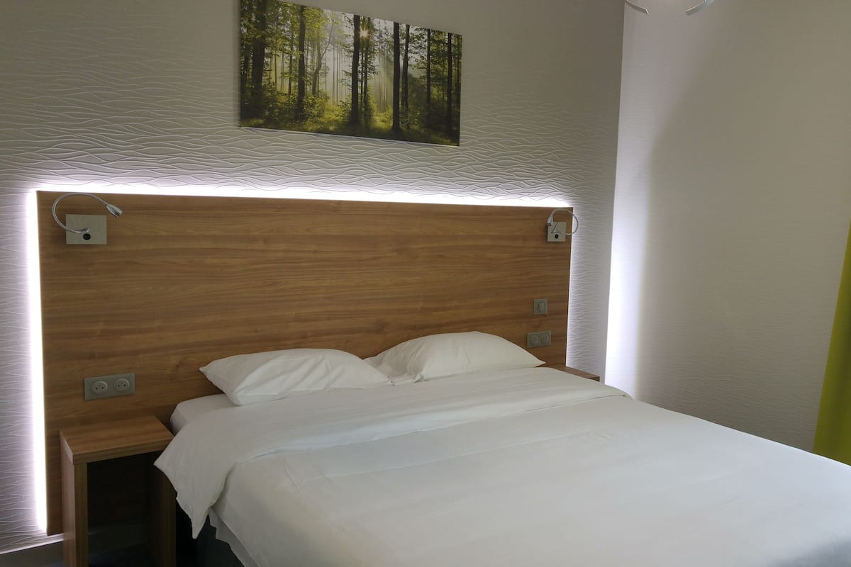 Una cama bien hecha con cabecera de madera, un cuadro de un bosque encima y una ventana con cortinas amarillas.