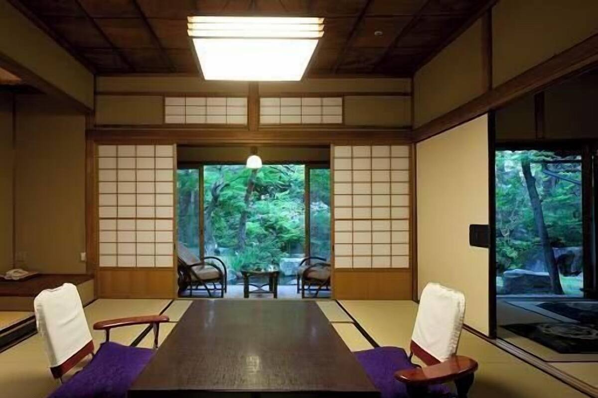 A traditional Japanese room with tatami flooring, a low table, and sliding doors leading to a view of greenery.