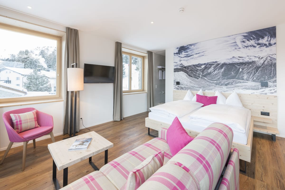 A modern hotel room with a large bed, a sofa, a small table, a pink chair, and a large window offering a view of the outside.