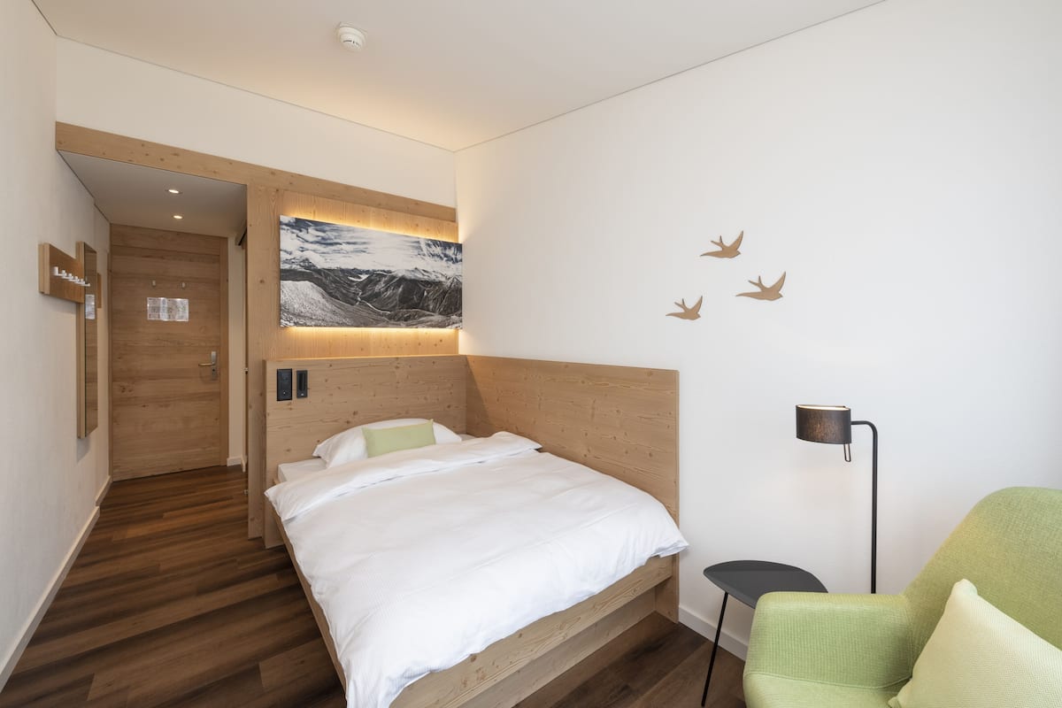 A modern hotel room with a wooden headboard, a large bed, a green armchair, and a wall-mounted lamp.