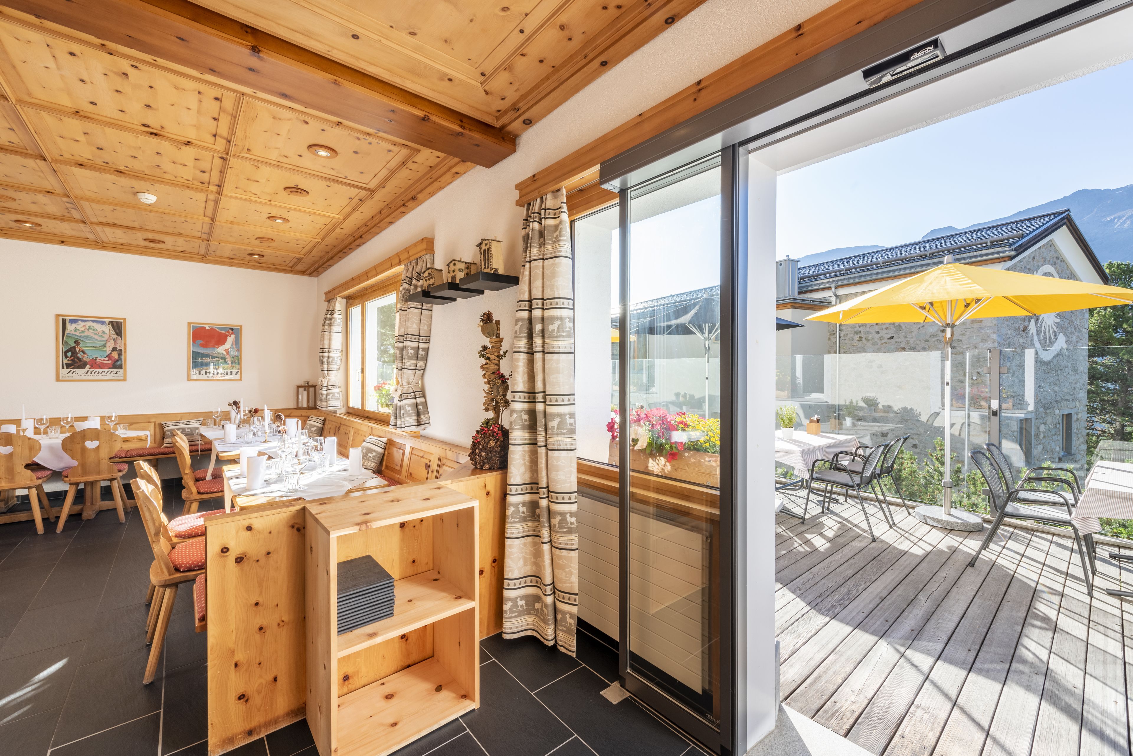 Ein Restaurant mit Holztischen, einer großen Schiebeglastür zu einer Terrasse und Blick auf Berge.