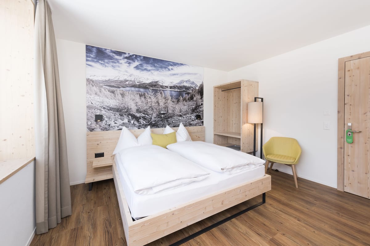 A bedroom with a wooden bed, a large landscape painting, a yellow chair, and a wooden wardrobe.