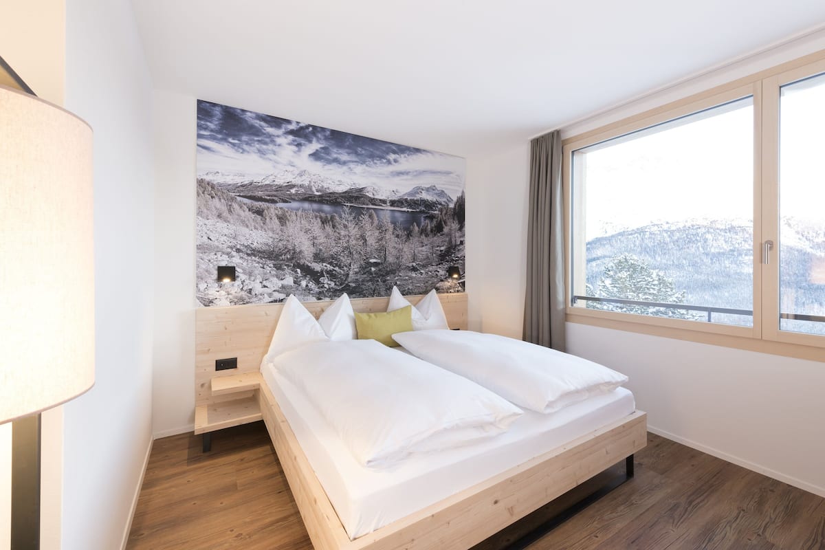 A bedroom with a wooden bed, a large window, and a mountain landscape painting on the wall.