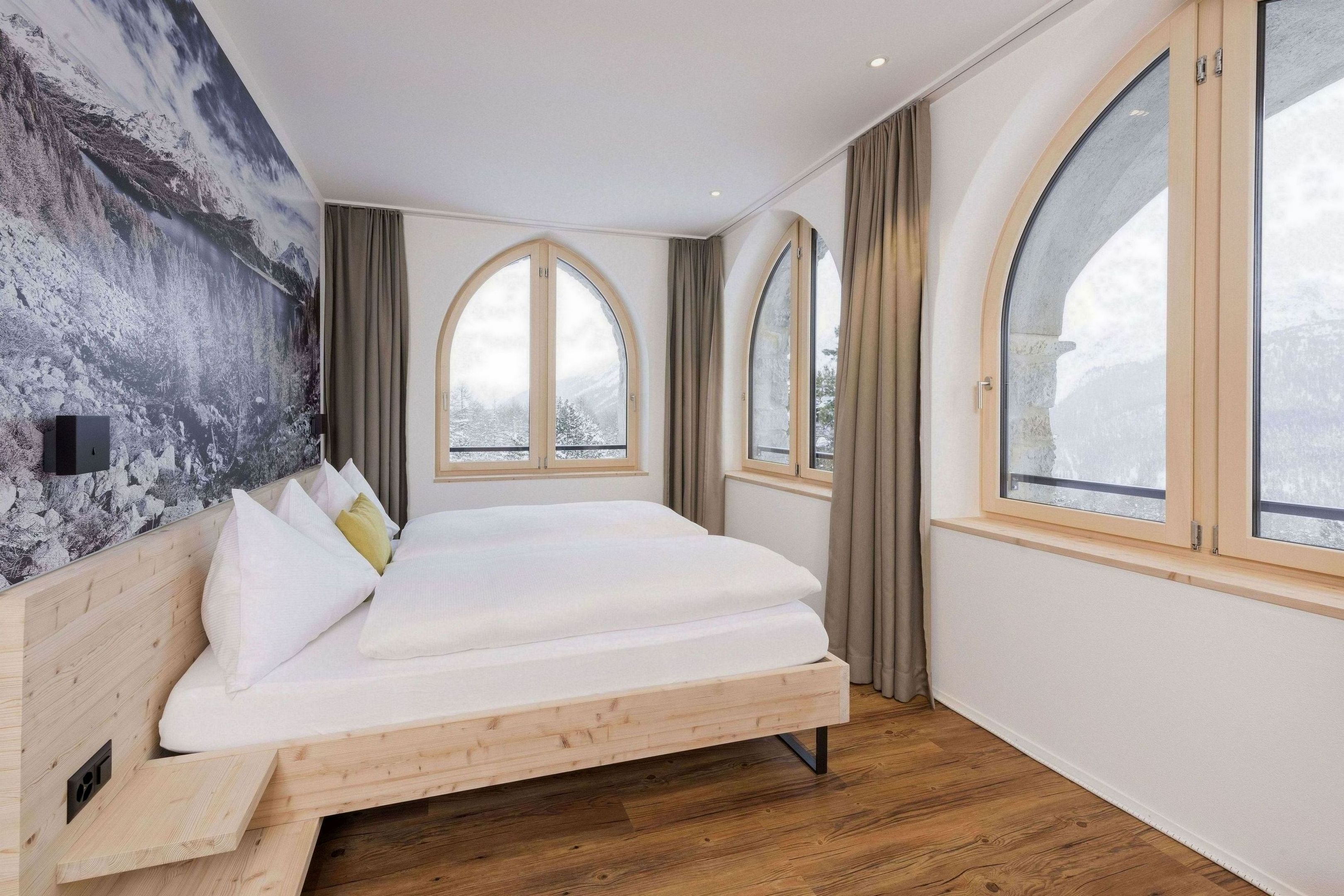 A modern bedroom with a wooden bed, large arched windows, and a mountain landscape mural on the wall.