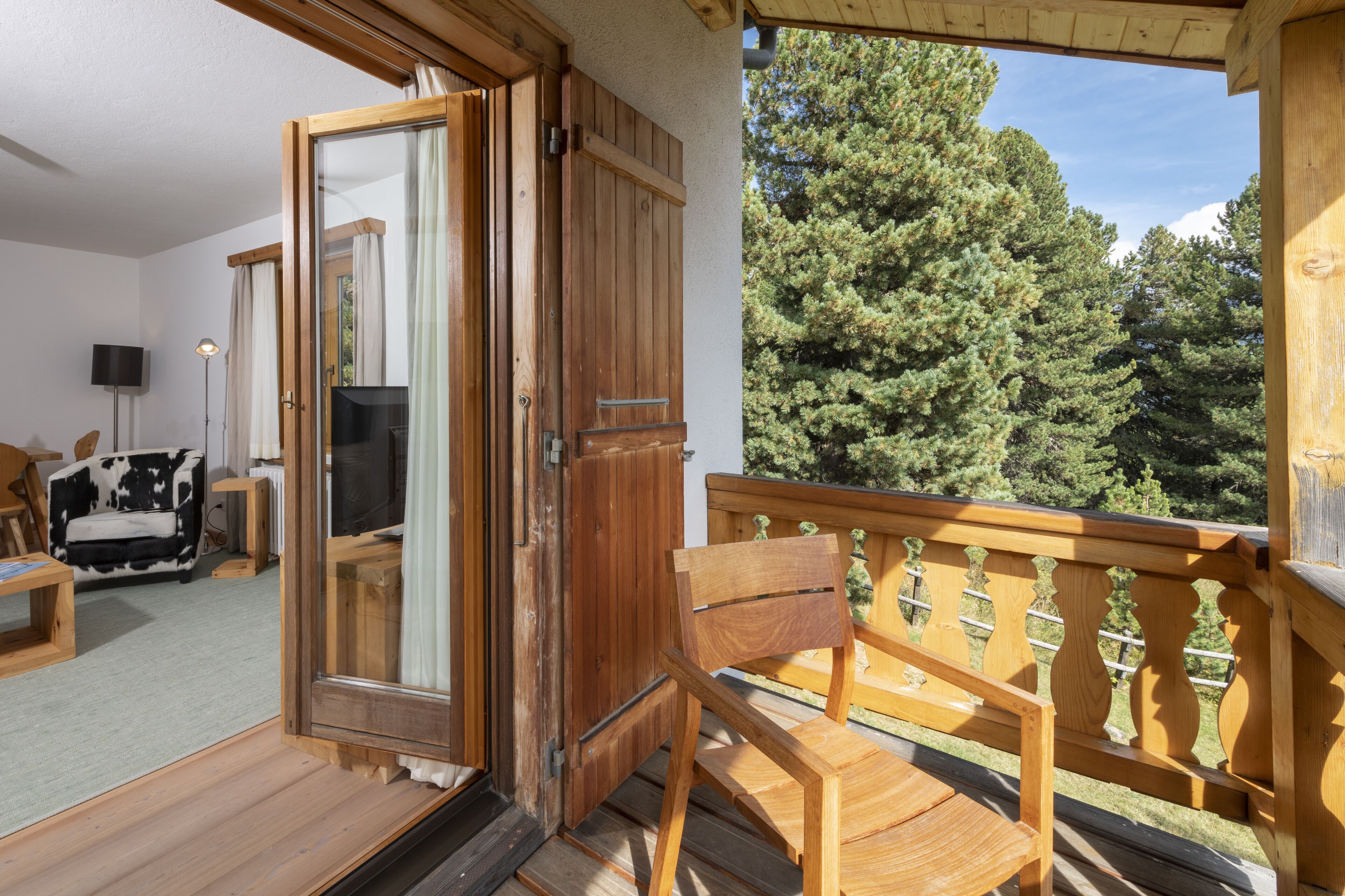 An open wooden door leading to a balcony with a view of trees. Inside, there is a wooden chair and a black and white sofa.