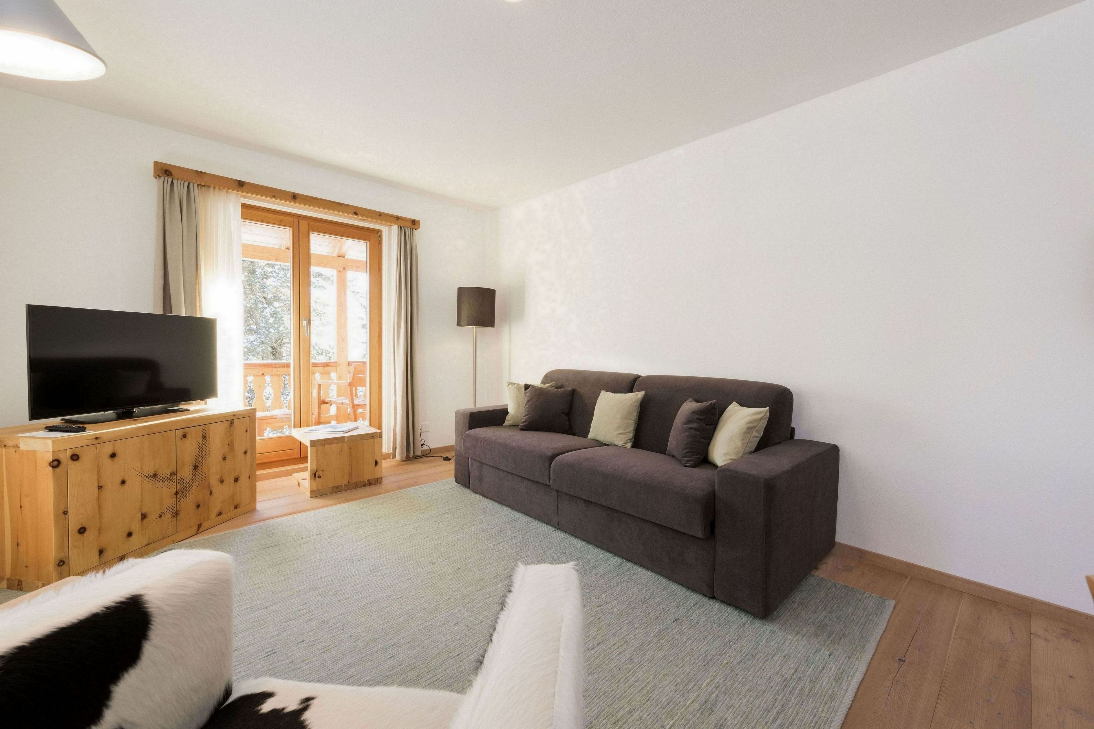 Un salon avec un grand téléviseur à écran plat posé sur un meuble en bois, un canapé marron foncé avec des coussins, un fauteuil blanc à poils longs et une grande fenêtre avec des rideaux.