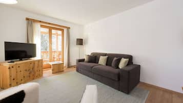 Un salon avec un grand téléviseur à écran plat posé sur un meuble en bois, un canapé marron foncé avec des coussins, un fauteuil blanc à poils longs et une grande fenêtre avec des rideaux.