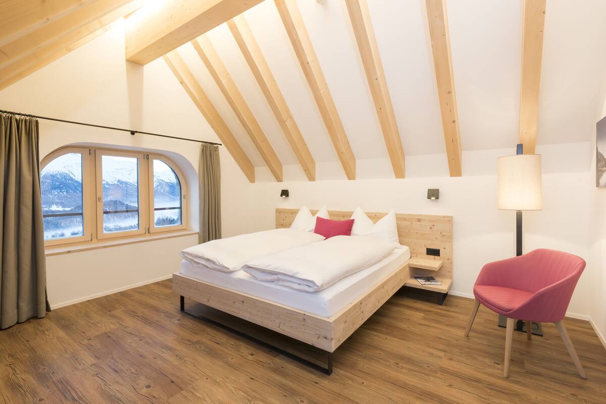 A bedroom with a wooden ceiling, a bed with white bedding, a red chair, and a window with a view of mountains.