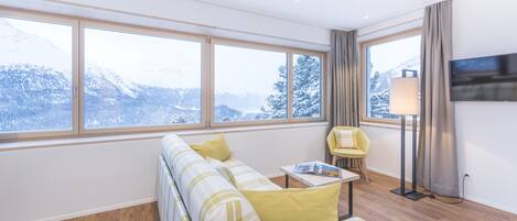 Un salón moderno con un sofá, una mesita, un sillón, una lámpara y un televisor. Amplios ventanales ofrecen vistas a montañas nevadas.