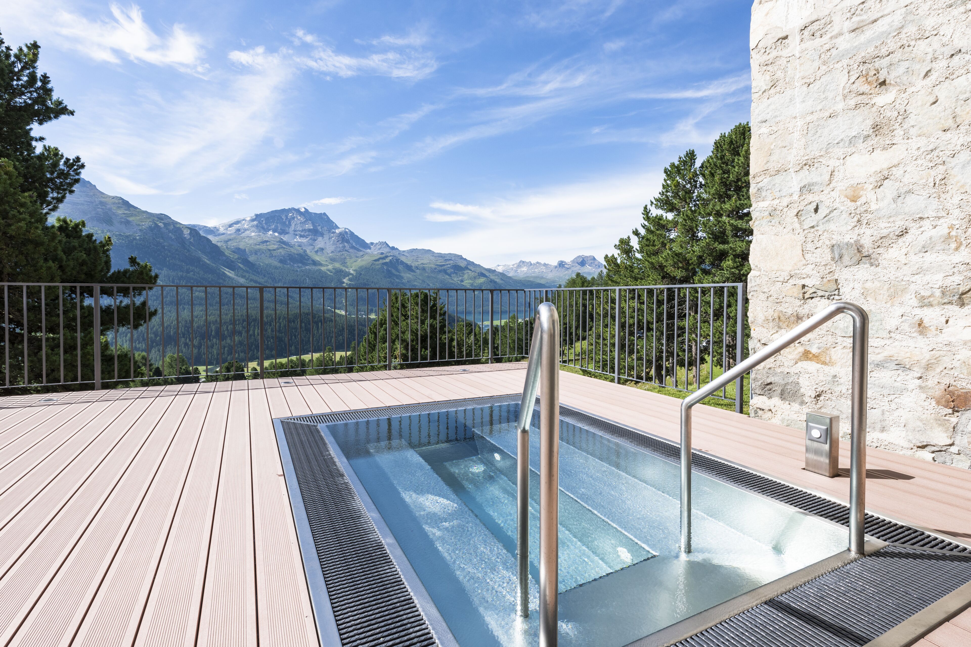 A rectangular outdoor pool with metal railings, surrounded by a wooden deck and a stone wall, overlooking a mountainous landscape.