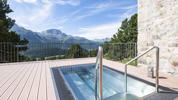 A rectangular outdoor pool with metal railings, surrounded by a wooden deck and a stone wall, overlooking a mountainous landscape.