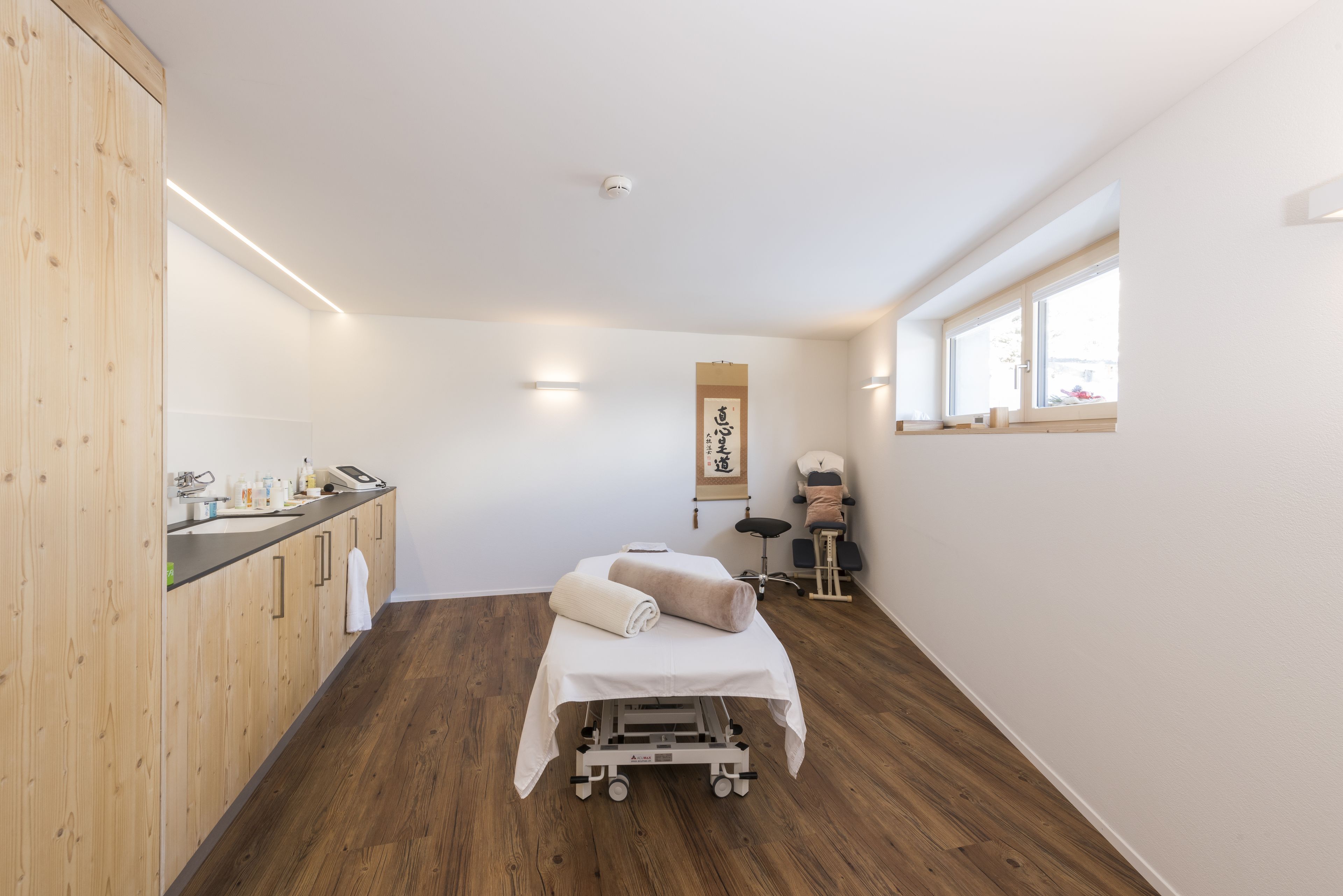 A room with a wooden floor, a bed with a white cover, a small table with a chair, and a window with a view.